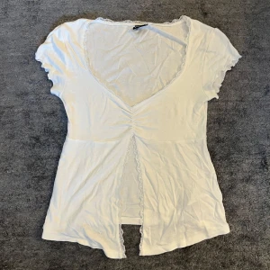 Vit spetsdetaljerad topp Gina Tricot - Supersöt vit topp från Gina Tricot med v-ringning och spetskant längs halsringningen. Toppen har korta ärmar, rynk framtill och en öppen slits med spetsdetalj. Perfekt för dig som gillar romantiska och feminina plagg.