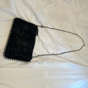 Svart axelväska med kedja och nitar - Snygg svart axelväska i mocka med silverfärgade kulnitar längs kanterna och en lång kedjerem. Väskan har dragkedja upptill och flera fack inuti, perfekt för mobilen och smågrejer. Ger en cool vibe till din outfit. Obs  knuten går enkelt att ta bort 
