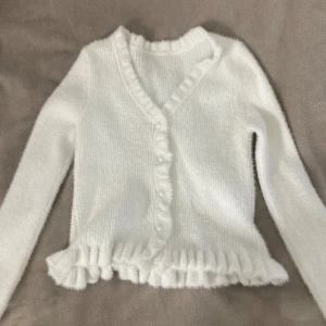 Vit fluffig kofta med volang  - Supermjuk och fluffig vit kofta från SHEIN i storlek S. Koftan har långa ärmar, volangkant nertill och längs knäppningen, samt pärlknappar framtill. Perfekt för lager-på-lager och ger en söt vibe till din outfit.