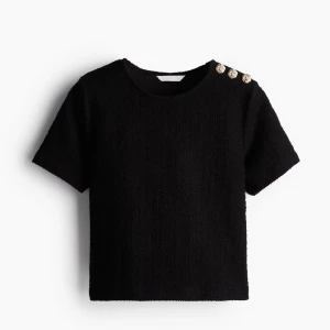 Svart kortärmad topp med guldknappar - Stilren svart topp i strukturerat tyg med korta ärmar och rund hals. Tre dekorativa guldknappar på ena axeln ger en lyxig touch. Använd enbart 1 gång, säljer pga den blivit för liten för mig. 