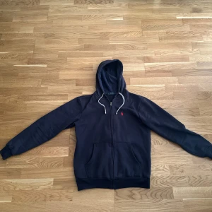Mörkblå hoodie från Polo Ralph Lauren - Snygg mörkblå hoodie från Polo Ralph Lauren med dragkedja, vit snörning i huvan och klassisk röd logga broderad på bröstet. Mjuk bomullskänsla och två stora fickor framtill. Perfekt för en chill och stilren look.