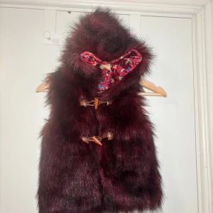 Vinröd pälsväst med huva från In Extenso - Säljer en vinröd pälsväst med huva från In Extenso. Västen har snygga träknappar framtill och ett färgglatt innerfoder med fågelmönster. Perfekt för dig som vill sticka ut med en mysig och cool look.