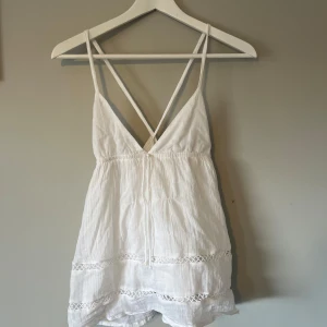 Vit linnetopp med spets från Gina Tricot - Supersöt vit linnetopp från Gina Tricot med tunna axelband som korsas i ryggen. Toppen har v-ringning, volangkant och fina spetsdetaljer längs nederdelen. Perfekt för varma dagar och ger en fräsch och luftig känsla.