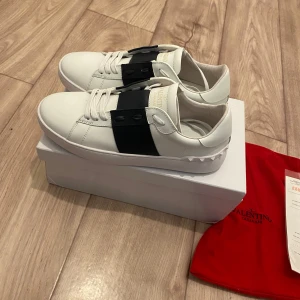 Valentino Garavani Open sneakers vit/svart - Snygga Valentino Garavani Open sneakers i vitt skinn med bred svart rem över vristen och vita snören. Klassisk rund tå och platt sula med ikoniska nitar baktill. Kommer med originalkartong och röd dustbag. Perfekta för dig som gillar exklusiva och stilrena sneakers.