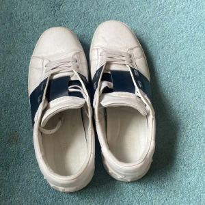 Valentino Garavani vita sneakers - Snygga vita sneakers från Valentino Garavani med bred mörkblå läderdetalj över vristen och ikoniska nitar på hälen. Skorna har snörning, rund tå och platt sula. Tillverkade i skinn för en lyxig känsla och stilren look.