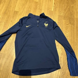 Frankrike långärmad Nike fotbollströja - Mörkblå långärmad fotbollströja från Nike med Frankrikes landslagsemblem och två stjärnor på bröstet. Guldiga detaljer och halv dragkedja framtill. Ärmarna har diskret mönster i blått. Perfekt för dig som älskar fotboll och vill representera Les Bleus.