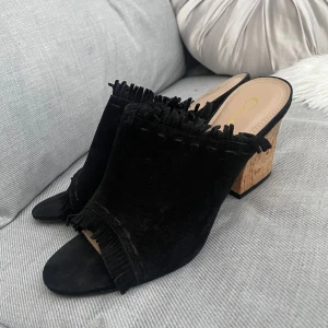 Svarta sandaler med korkklack från Gacel - Svarta sandaler i mocka med öppen tå och fransdetaljer längs kanterna. Snygg korkklack som ger en cool kontrast till det svarta. Perfekta för dig som gillar statementskor med unik touch.