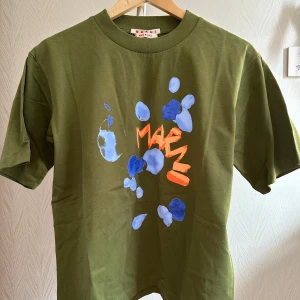 Grön t-shirt med print från Marni - Snygg grön t-shirt från Marni med coolt blått och orange print framtill. Oversized fit och klassisk rund hals. Tillverkad i 100% bomull för en skön känsla. Perfekt för dig som gillar statement-plagg och unika designer.