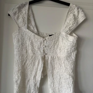 Vit topp från Gina Tricot - Supersöt vit topp från Gina Tricot med smockad struktur och breda axelband. Toppen har knappar framtill och är tillverkad i ett lätt, krinklat tyg som ger en cool och avslappnad vibe. Perfekt för varma dagar och sommarhäng.