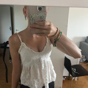 Vit broderad linnetopp med spets - Supersöt vit linnetopp med tunna axelband och broderade blommor. Toppen har volangkant vid urringningen och är lätt utsvängd nertill. Perfekt för varma dagar och ger en romantisk vibe. Materialet känns luftigt och toppen är ärmlös.