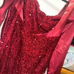 Vinröd glittrig långklänning L - Säljer en vinröd långklänning med långa ärmar och massor av paljetter som ger ett riktigt glittrigt intryck. Klänningen har en snygg draperad urringning och är tillverkad i ett satinliknande material med paljettöverdrag framtill. Perfekt för bal eller fest!