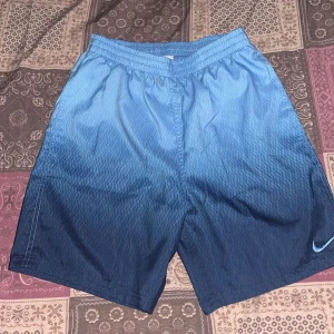 Nike blå ombre shorts - Snygga blå shorts från Nike med ombre-effekt som går från ljusblått upptill till mörkblått nedtill. Elastisk midja för skön passform och diskret Nike-logga på benet. Perfekta för sommarens alla aktiviteter.
