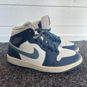 Nike Air Jordan 1 Mid blå/vit - Nike Air Jordan 1 Mid i blått och vitt med klassisk Jordan-logga på sidan. Skorna har snörning, perforerad tå och ikonisk swoosh. Ovandel i skinn och textil, mellanhög modell som ger en clean och sportig vibe.
