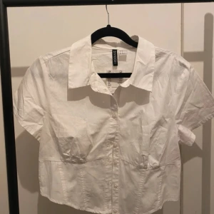 Vit croppad skjorta från H&M Divided - Snygg vit croppad skjorta från H&M Divided med korta ärmar, klassisk krage och knappar framtill. Skjortan har smockad detalj baktill för en cool passform. Perfekt till jeans eller kjol för en fräsch look. Oanvänd med prislapp.