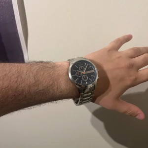 Armani Exchange silverfärgad klocka - Stilren klocka från Armani Exchange med silverfärgad länk i metall och mörkblå urtavla. Klockan har tre mindre urtavlor, orange detaljer och tydliga index. Perfekt accessoar för dig som gillar en modern och sportig look. Kommer med lådan nypris 2500kr. Klockan är helt ny köpte för ett bröllop som inte blev av 