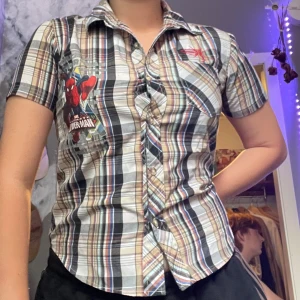 Baby skjorta Rutig Spider-Man🪩 - Kortärmad rutig skjorta med färger i beige, svart, blått och rött. Stor Spider-Man-tryck på bröstet och broderad text på ryggen. Klassisk krage, knappar framtill och bröstficka. Perfekt för dig som vill sticka ut med Marvel-vibe.