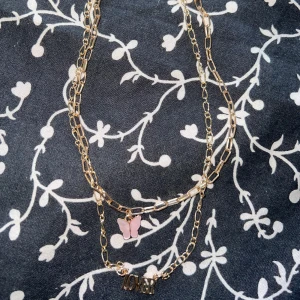 Gulfärgad halsband med fjäril och text - Dubbelkedjat halsband i guldtonad metall med chunky länkar. Ena kedjan har ett hänge med rosa fjäril och den andra har ett hänge med texten 'LOVER'. Perfekt statement-piece för att lyfta din outfit. Öppnad förpackning. 