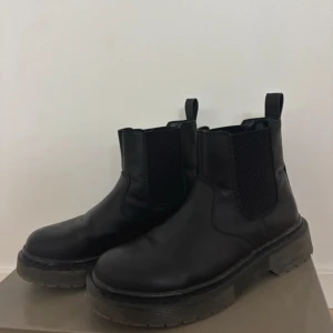 Svarta chelsea boots i skinn - Chunky svarta chelsea boots med bred resår på sidorna och grov platt sula. Tillverkade i skinn med rund tå och dragflikar både fram och bak. Perfekta för höst och vinter när du vill ha en stilren men edgy look.