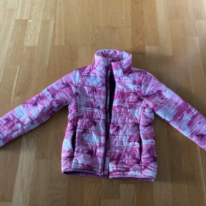 Rosa och vit pufferjacka från Everest - Säljer en färgstark pufferjacka från Everest i storlek 122/128. Jackan har ett unikt rosa och vitt mönster, avtagbar huva och dragkedja framtill. Perfekt för dig som gillar att sticka ut och vill ha en varm jacka med cool vibe.