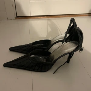 Svarta spetsiga pumps från Qimeluq - Snygga svarta pumps med spetsig tå och smal klack. Skorna har ett glansigt material med dekorativa sömmar och ett silvrigt innersula. Rem runt vristen med spänne för extra support. Perfekta för dig som vill sticka ut med en elegant och edgy look.