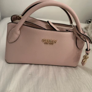 Ljusrosa handväska från Guess - Superfin ljusrosa handväska från Guess med guldfärgade detaljer och logga framtill. Väskan har både handtag och justerbar axelrem, samt dragkedja upptill. Tillverkad i strukturerad syntet med flera fack inuti för smart organisering.