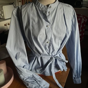 Skjorta/blus med knyte - Snygg ljusblå blus med ståkrage, pärlor som knappar framtill och knytband i midjan som ger en markerad siluett. Långa ärmar med knäppning vid ärmslut. Perfekt för dig som gillar stilrena och trendiga plagg. Går som en M i storleken
