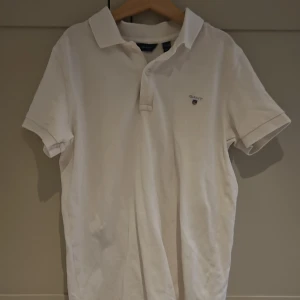 Vit pikétröja från GANT - Klassisk vit pikétröja från GANT med korta ärmar och krage. Tröjan har broderad logga på bröstet och knäppning framtill. Perfekt för en clean och stilren look. Tillverkad i mjuk bomull som känns skön mot huden.