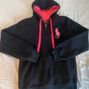 Svart/Rosa hoodie från Polo Ralph Lauren Storlek XL - Svart hoodie från Polo Ralph Lauren med dragkedja, rosa detaljer och stor rosa logga på bröstet. Snören i rosa, samt broderad siffra 3 på ena ärmen. Perfekt för dig som gillar sportig och stilren streetwear. På etiketten står det storlek XL, men passar även bra fast lite mer baggy om du har en mindre storlek (Jag är vanligtsvis XS/S för referens).