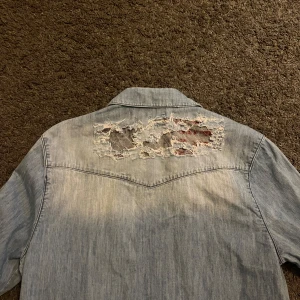 Ljusblå jeansskjorta med slitningar - Säljer en ljusblå jeansskjorta från Adrian hammond med coola detaljer på axlar och rygg. Skjortan saknar en knapp (syns på sista bilden) men är annars i bra skick.