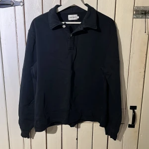 Svart pikétröja från East West - Klassisk svart pikétröja från East West i storlek L. Tröjan har lång ärm, krage och knäppning framtill. Perfekt för en clean och stilren look. Tillverkad i mjuk bomull som känns skön mot huden. Sitter som en M