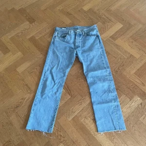 Levi's 501 ljusblå jeans W32 L30 - Klassiska Levi's 501 jeans i ljusblå denim med raka ben och råa, fransiga benslut. Femficksmodell med knappgylf och ikonisk läderpatch bak i midjan. Perfekta för en avslappnad och tidlös look.