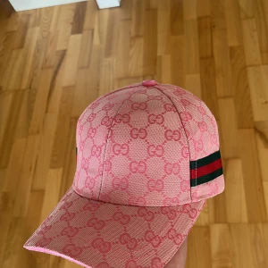 Rosa Gucci keps med GG-mönster - Snygg rosa keps från Gucci med ikoniskt GG-mönster över hela kepsen. Kepsen har klassisk böjd skärm, justerbart spänne bak i rosa läder och en grön-röd rand på sidan. Tillverkad i bomull och detaljer i skinn. Perfekt för dig som vill sticka ut.