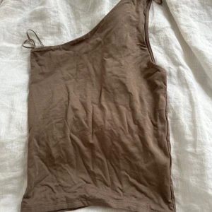 Brun oneshoulder topp  - Snygg brun oneshoulder topp med smal axelrem på ena sidan. Enkel och stilren design i mjuk bomull, perfekt för att skapa en trendig look. Passar till både jeans och kjol för en cool vibe.