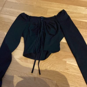 Svart croppad topp från SHEIN - Trendig svart croppad topp från SHEIN med långa ärmar och snygg snörning framtill. Offshoulder-modell som ger en cool vibe och passar perfekt till höga jeans eller kjol. Tunn och stretchig, perfekt för att skapa en edgy look.