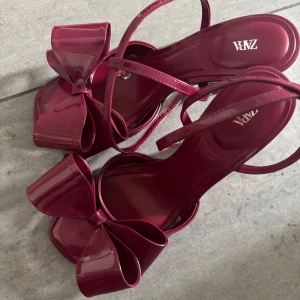 Vinröda klackpumps med rosett från Zara - Snygga vinröda pumps från Zara med hög klack och stora glansiga rosetter framtill. Skorna har smala remmar och öppen tå, vilket ger en elegant och trendig look. Perfekta för dig som vill sticka ut med en färgstark detalj. Dem är även helt oanvända