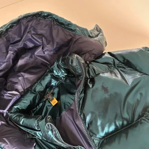 Mörkgrön pufferjacka från Polo Ralph Lauren - Säljer en mörkgrön, glansig pufferjacka från Polo Ralph Lauren. Jackan har hög krage, avtagbar huva och dragkedja framtill. Klassisk broderad logga på bröstet och två fickor med dragkedja. Perfekt för kalla dagar och riktigt snygg streetstil.