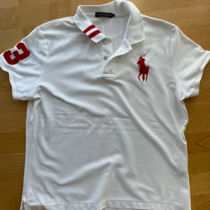 Vit pikétröja från Ralph Lauren - Vit pikétröja från Ralph Lauren med röd broderad logga på bröstet och röda ränder på kragen. Klassisk pikékrage, korta ärmar och knappslå framtill. Stor röd siffra 3 på ena ärmen. Tillverkad i bomull för en skön och luftig känsla. Tröjan är i syl XL men är liten i storleken. Jag är normalt en S/M men har som sagt haft den här i XL. 