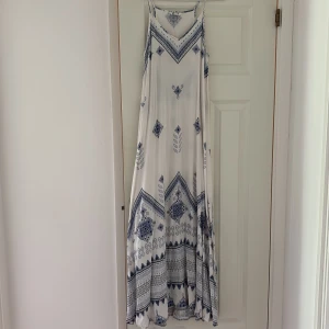 Vit och blå långklänning med mönster - Luftig långklänning i vitt med blått bohemiskt mönster. Klänningen har smala axelband och ett flowy fall, perfekt för varma dagar. Snygg och avslappnad stil med fina detaljer i mönstret.