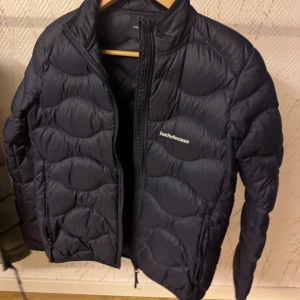 Mörkblå Peak Performance pufferjacka - Snygg mörkblå pufferjacka från Peak Performance med vågigt quiltad design och diskret logga på bröstet. Jackan har dragkedja framtill och är tillverkad i lätt polyester, perfekt för kyliga dagar. Passformen är normal och jackan har långa ärmar. Passar storlek S
