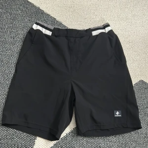 Svarta shorts Cross Sportswear  - Svarta shorts från Cross Sportswear med bred elastisk midja i grått, vitt och svart randigt mönster. Modellen har normal passform och är tillverkad i ett lätt syntetmaterial, perfekt för sportiga aktiviteter. Diskret logga framtill och snörning i midjan.