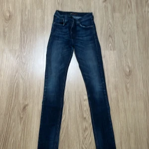 Mörkblå skinny jeans från Nudie Jeans - Snygga mörkblå jeans från Nudie Jeans med skinny passform och klassisk femficksdesign. Jeansen har coola slitningar, bruna detaljer och Nudie-logga på bakfickan. Perfekt för dig som gillar en smal siluett och stilren look.