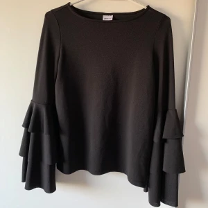 Svart topp med volangärmar från Ginatricot  - Svart långärmad topp från Ginatricot med riktigt snygga volangärmar i flera lager. Rund halsringning och loose passform som ger en chill vibe. Perfekt om du vill sticka ut lite extra med coola detaljer på ärmarna.