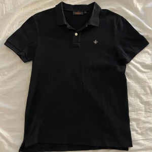 Svart pikétröja Morris Stockholm  - Svart pikétröja från Morris Stockholm i storlek S. Klassisk design med krage, två knappar och broderad logga på bröstet. Tillverkad i mjuk bomull som är skön mot huden. Perfekt för dig som gillar stilrena och enkla plagg.