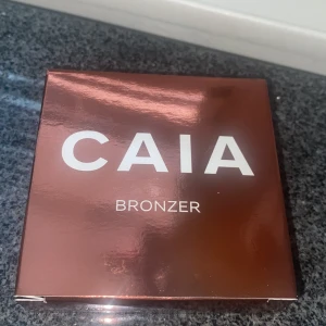 CAIA Bronzer i lyxig förpackning - Snygg bronzer från CAIA i en glansig, kopparfärgad fyrkantig dosa med spegel. Pudret är varmt brunt med en lätt skimrig finish och har cirkulärt mönster med CAIA-loggan i mitten. Perfekt för att ge ansiktet en solkysst look.