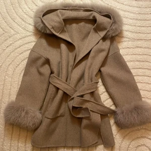 Beige kappa med pälsdetaljer och huva - Snygg beige kappa med fluffiga pälsdetaljer på huva och ärmslut. Jackan har knytbälte i midjan, stora fickor framtill och en oversized huva. Materialet är ull och päls, perfekt för kalla dagar. Orginalpris - 2999 Nypris - 1500 