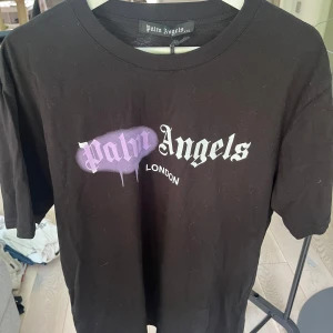 Svart Palm Angels t-shirt London - Svart t-shirt från Palm Angels med tryck på bröstet där 'Palm' är sprayat i lila och 'Angels' står i vit gotisk stil. Under står 'London' i mindre vit text. Klassisk rund hals och korta ärmar. Perfekt för dig som gillar streetwear och coola prints.