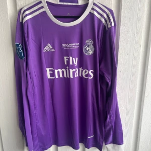 Real Madrid Ronaldo 7 lila långärmad tröja - Säljer en officiell Real Madrid fotbollströja från Adidas, Champions League Final Cardiff 2017 edition. Tröjan är lila med vita detaljer, långärmad och har Ronaldo 7 tryckt på ryggen. Materialet är lätt och andas, perfekt för match eller träning.