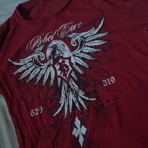 Röd Rebel Spirit t-shirt med örntryck - Snygg röd t-shirt från Rebel Spirit med stort vitt och svart örntryck på framsidan, coola siffror och texten 'Rebel Spirit' upptill. Klassisk passform och rund hals. Perfekt för dig som vill sticka ut med en unik design.