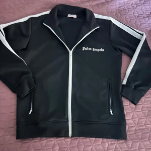 Svart track jacket från Palm Angels - Palm Angels svart track jacket med vita ränder längs ärmarna och vit dragkedja. Jackan har ståkrage, två fickor med dragkedja och logotypen tryckt på bröstet. Materialet är polyester och modellen är loose fit med långärmade ärmar.