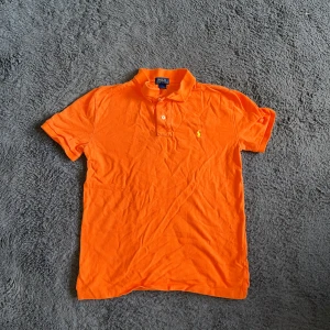 Ralph Lauren piké  - Säljer en fräsch orange pikétröja från Polo Ralph Lauren med klassisk krage och två knappar framtill. Tröjan har korta ärmar och den ikoniska lilla broderade loggan i gult på bröstet. Materialet är mjuk bomull som andas och passar perfekt till sommaren. Storlek L 14-16 år vilket bör motsvara XS men KOLLA MÅTTEN!